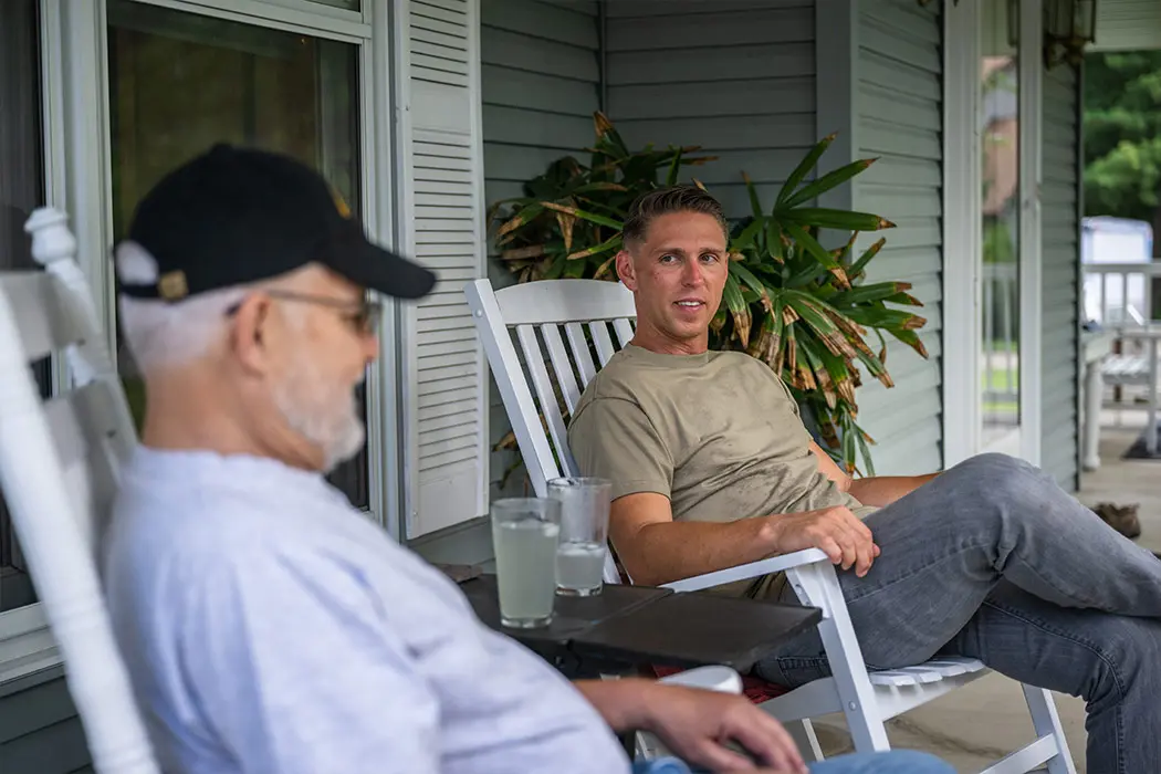 Young man speaking to his dad about veteran hospice care on the porch sitting in rocking chairs.